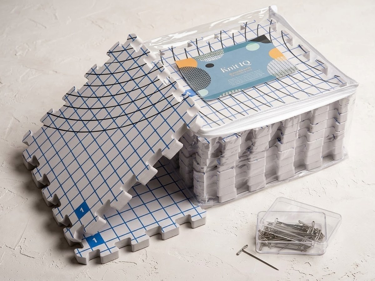 A set of KnitIQ interlocking foam blocking mats printed with a blue grid, stacked and packed into a clear zippered storage bag beside a box of stainless T-pins and a loose pin.