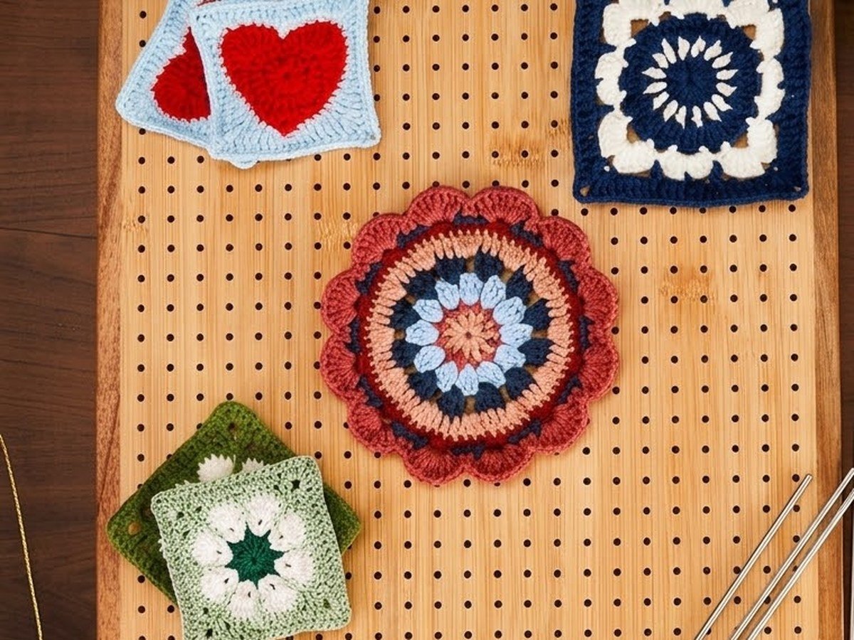 Four crochet motifs of different shapes — two hearts, a floral square, a round mandala and a granny square — pinned at various points across the blocking board’s hole grid.